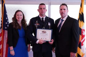 MVA Administrator Chrissy Nizer stands to the left of TSS III Designee Brandon Wright, who holds a certificate. Law Enforcement Liaison Tom Lubinski stands to his right. The three are framed by an American flag on the left and a Maryland flag on the right. 